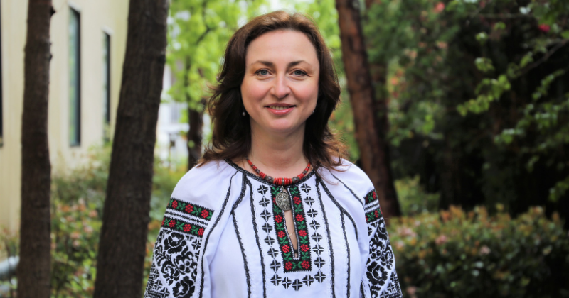 Professor Iryna Zaverukha in a traditional Ukrainian embroidered blouse standing in front of green foliage 