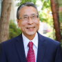 Professor Dennis Yokoyama headshot in dark suit and red tie in front of greenery and trees