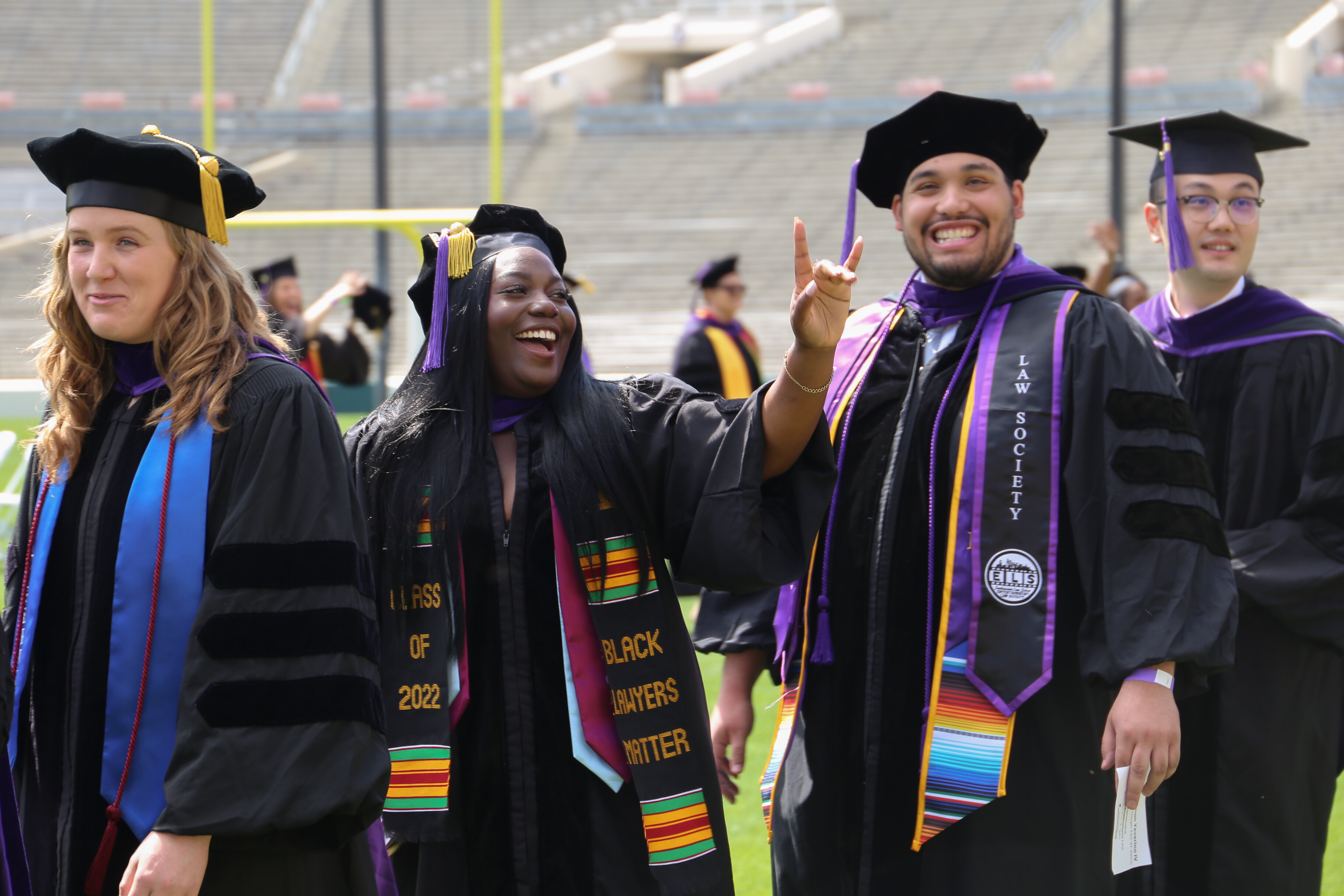 Four students in commencement regalia walking across the lawn smiling