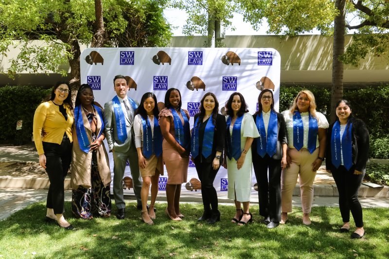 Jeff Martin in a grey suit and Public Service Program sash standing in a line in front of a Southwestern Law School step and repeat backdrop with Michelle Takagishi-Almeida and the other graduating student award recipients at the Public Service Program luncheon