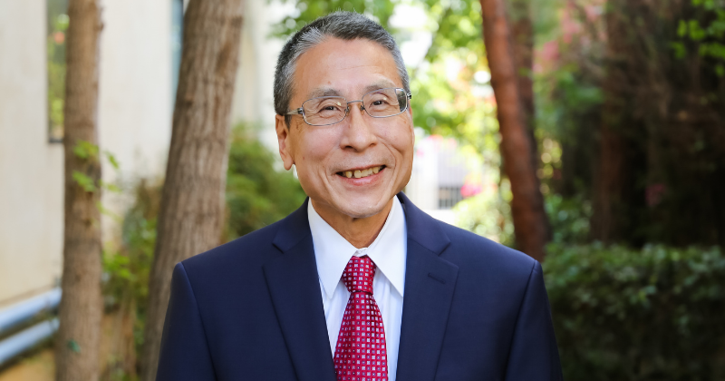 Professor Dennis Yokoyama headshot in dark suit and red tie in front of greenery and trees